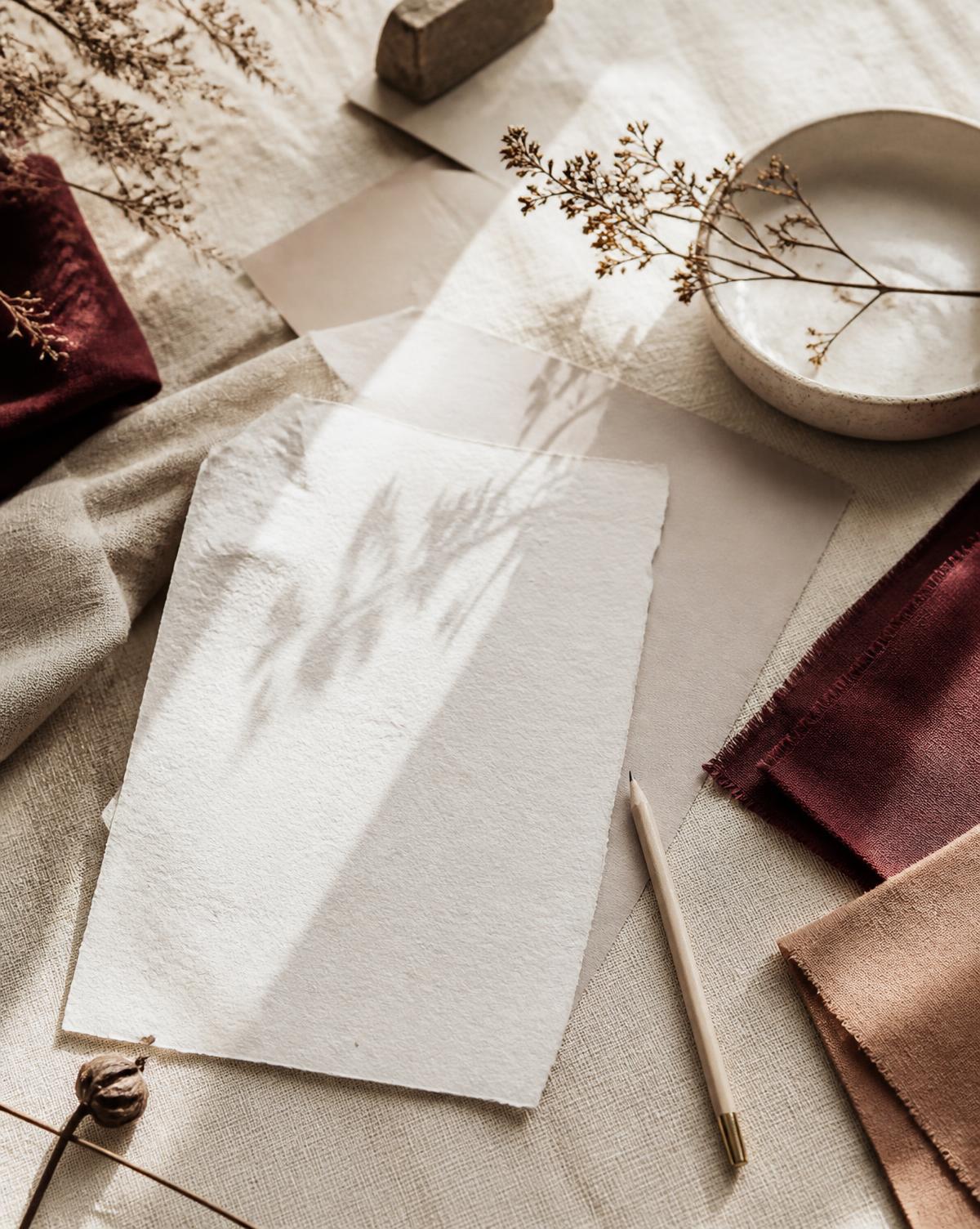 Flat lay editoriale di carta, tessuti in bordeaux e beige, ceramica e fiori secchi su tavolo in lino