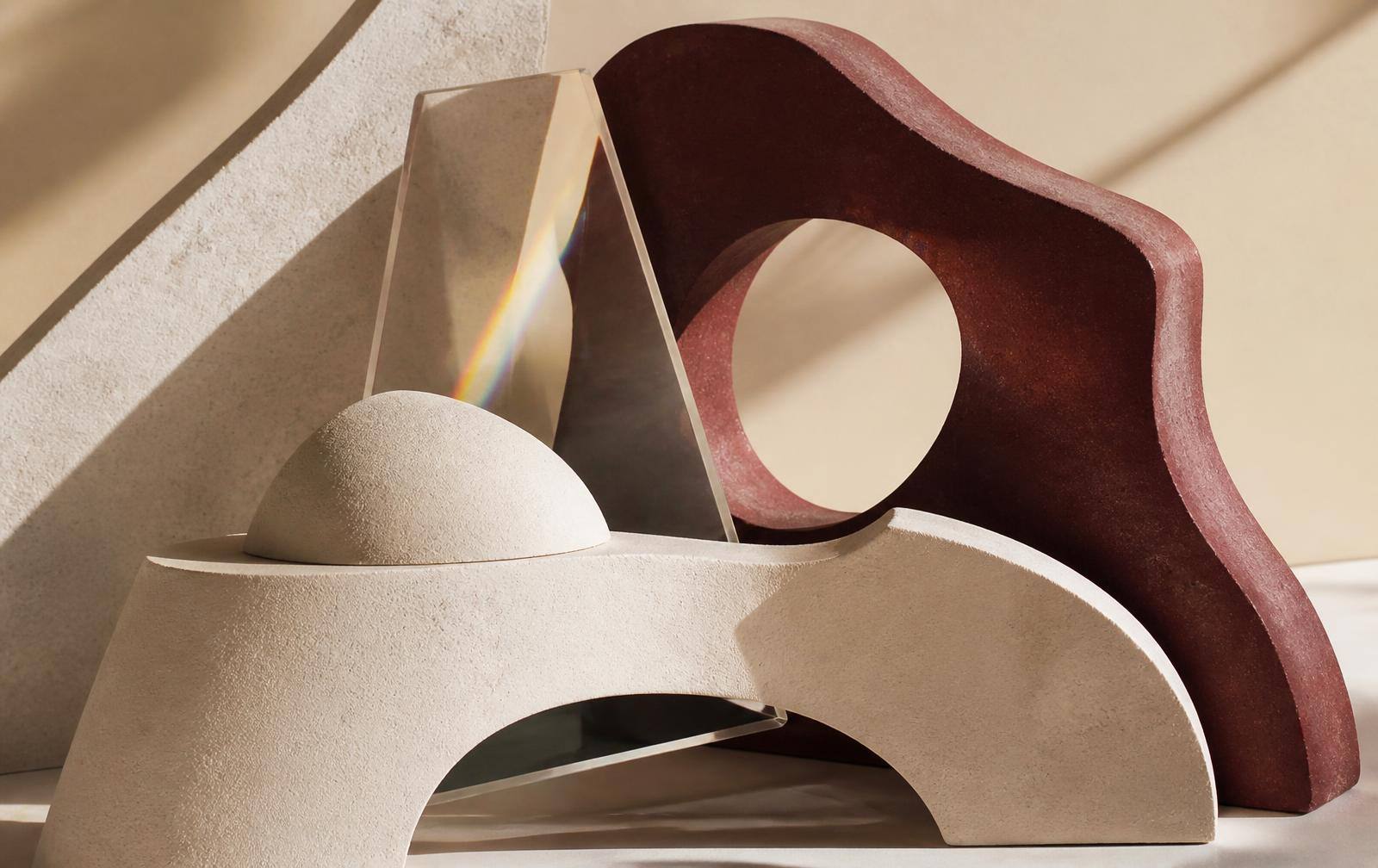 Forme scultoree in gesso crema e ceramica bordeaux con prisma di vetro e ombre morbide